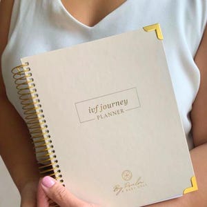 May include: A beige spiral-bound planner with gold accents, featuring the text "ivf journey PLANNER." The planner has gold corner protectors and a gold spiral binding. The bottom of the planner has the text "By Paola & Baby Dust."