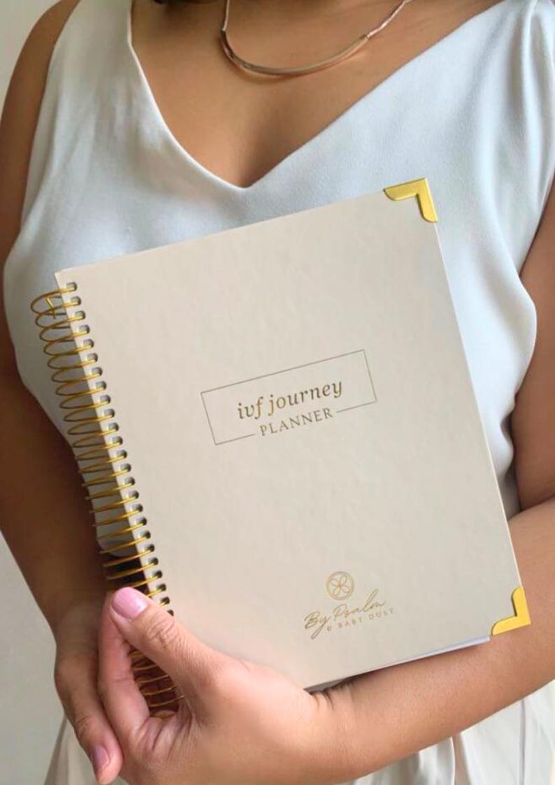 May include: A beige spiral-bound planner with gold accents, featuring the text "ivf journey PLANNER." The planner has gold corner protectors and a gold spiral binding. The bottom of the planner has the text "By Paola & Baby Dust."