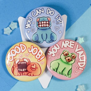 May include: Three round buttons with colorful cartoon frog designs and text. The first button is light blue with the text "You Can Do It!" and a blue frog with a toothy grin. The second button is pink with the text "You Are Valid!" and a green frog with large eyes. The third button is yellow with the text "Good Job!" and a red and orange monster with a toothy grin.