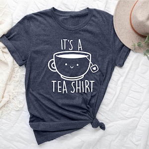 May include: Dark gray t-shirt with the text "It's a Tea Shirt" and a smiling teacup graphic. The graphic is white and features a teabag. The shirt is knotted at the bottom.