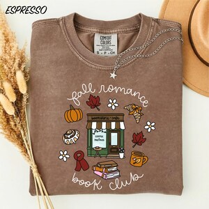 May include: A brown crewneck t-shirt featuring a graphic print with the text "fall romance book club" and "bookstore cafe coffee pastries". The design includes illustrations of a bookstore, books, a pumpkin, leaves, flowers, a scarf, and a mug.