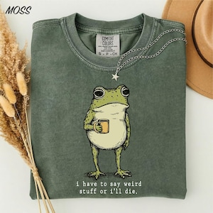 May include: A moss green crew neck t-shirt featuring a graphic illustration of a green frog holding a small yellow mug. Below the frog, the text reads: I have to say weird stuff or i'll die. The shirt has a vintage-style wash and a tag reading Comfort Colors.