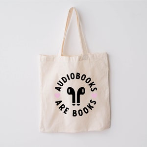 K&ouml;nnte beinhalten: Beige Canvas-Tragetasche mit schwarzem Text und einer Grafik von Ohrh&ouml;rern. Der Text lautet "AUDIOBOOKS ARE BOOKS" in einem kreisf&ouml;rmigen Design, mit zwei rosa Herzen. Die Tasche hat zwei Griffe.