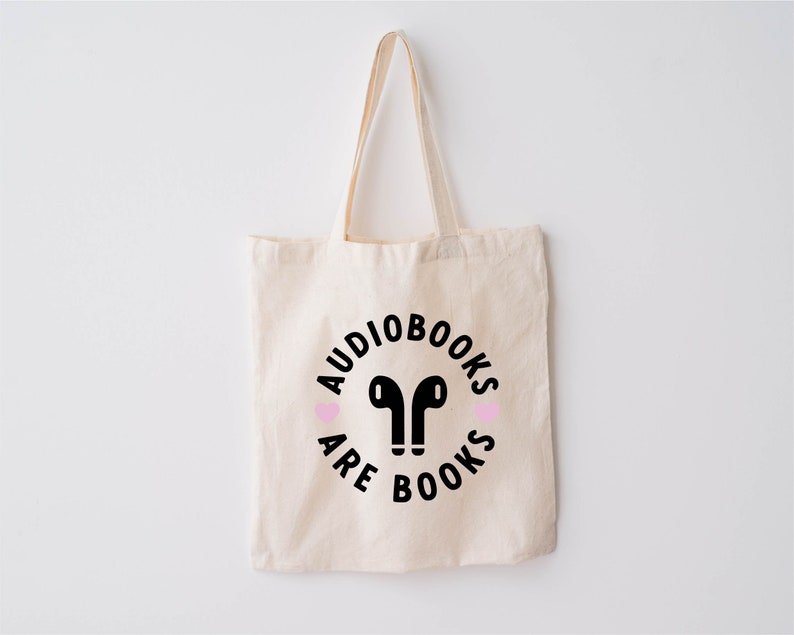 K&ouml;nnte beinhalten: Beige Canvas-Tragetasche mit schwarzem Text und einer Grafik von Ohrh&ouml;rern. Der Text lautet "AUDIOBOOKS ARE BOOKS" in einem kreisf&ouml;rmigen Design, mit zwei rosa Herzen. Die Tasche hat zwei Griffe.