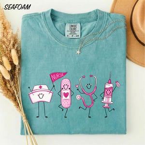 A turquoise t-shirt with a graphic design depicting four cartoon nurse characters, including a nurse's hat, stethoscope, bandage, and syringe, along with the text "NURSE" and "CONFIDENCE COLOR".