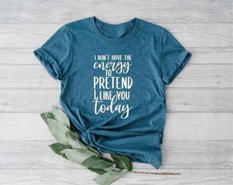 May include: A teal-colored t-shirt with the text "I don't have the energy to pretend I like you today" in white script. The shirt is knotted at the bottom and has rolled-up sleeves. Green leaves are placed near the shirt.
