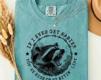 Puede incluir: Una camiseta verde espuma de mar con un gráfico negro de un mapache gruñendo encerrado en un círculo de alambre de púas. El texto dice IF I EVER GET RABIES YOU'RE HIGH ON MY BITIN' LIST. La camiseta tiene una etiqueta Comfort Colors.