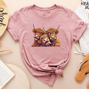 a pink t-shirt with a graphic of three cows, two of which are making faces, and the third one is sticking out its tongue. The t-shirt is displayed on a table with various items, including a hat, a book, and a pair of sandals.