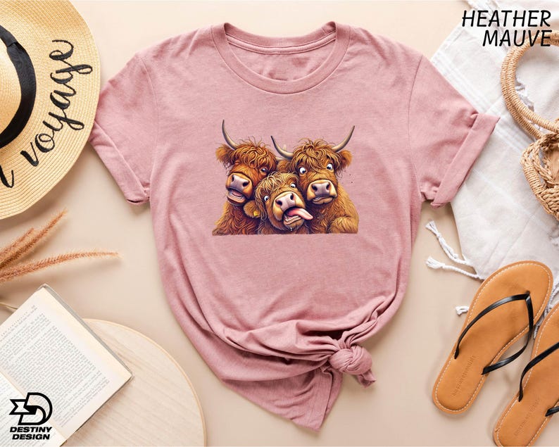 a pink t-shirt with a graphic of three cows, two of which are making faces, and the third one is sticking out its tongue. The t-shirt is displayed on a table with various items, including a hat, a book, and a pair of sandals.