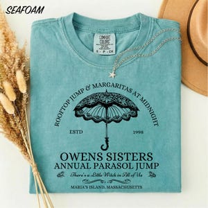 May include: A seafoam green short-sleeve t-shirt featuring a black graphic of a lace parasol. The text reads: ROOFTOP JUMP & MARGARITAS AT MIDNIGHT, ESTD 1998, OWENS SISTERS ANNUAL PARASOL JUMP, There's a Little Witch in All of Us, MARIA'S, MASSACHUSETTS.