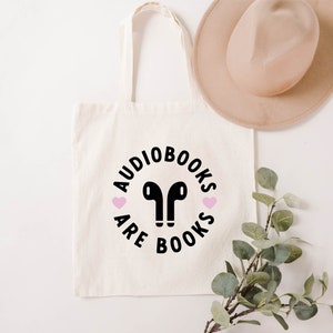 K&ouml;nnte beinhalten: Cremefarbene Tragetasche mit schwarzem Text "Audiobooks are Books" in einem kreisf&ouml;rmigen Design, mit einem Paar schwarzer Ohrh&ouml;rer in der Mitte und zwei rosa Herzen. Ein beiger Hut und eine gr&uuml;ne Pflanze liegen neben der Tasche.