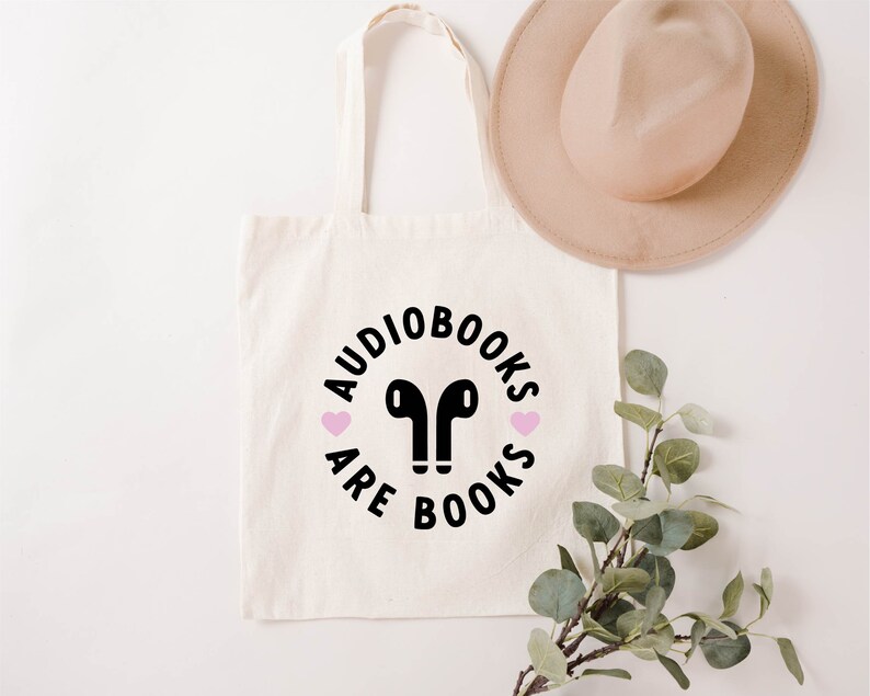 K&ouml;nnte beinhalten: Cremefarbene Tragetasche mit schwarzem Text "Audiobooks are Books" in einem kreisf&ouml;rmigen Design, mit einem Paar schwarzer Ohrh&ouml;rer in der Mitte und zwei rosa Herzen. Ein beiger Hut und eine gr&uuml;ne Pflanze liegen neben der Tasche.