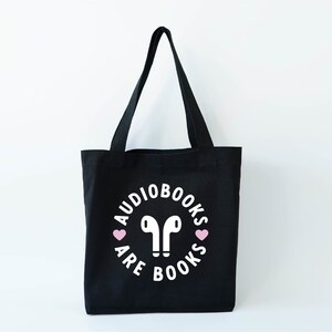 K&ouml;nnte beinhalten: Schwarze Tragetasche mit wei&szlig;em Text und rosa Herzen. Der Text lautet "Audiobooks are books" in einem kreisf&ouml;rmigen Design um ein Paar wei&szlig;er Ohrh&ouml;rer. Die Tasche hat zwei schwarze Griffe und steht vor einem wei&szlig;en Hintergrund.