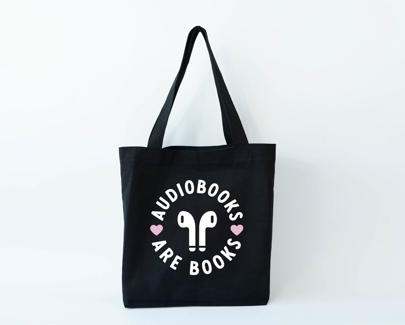 K&ouml;nnte beinhalten: Schwarze Tragetasche mit wei&szlig;em Text und rosa Herzen. Der Text lautet "Audiobooks are books" in einem kreisf&ouml;rmigen Design um ein Paar wei&szlig;er Ohrh&ouml;rer. Die Tasche hat zwei schwarze Griffe und steht vor einem wei&szlig;en Hintergrund.