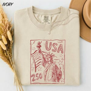 A beige t-shirt with a red graphic design depicting the Statue of Liberty and the text "USA 250" printed on it, along with a necklace and a hat.