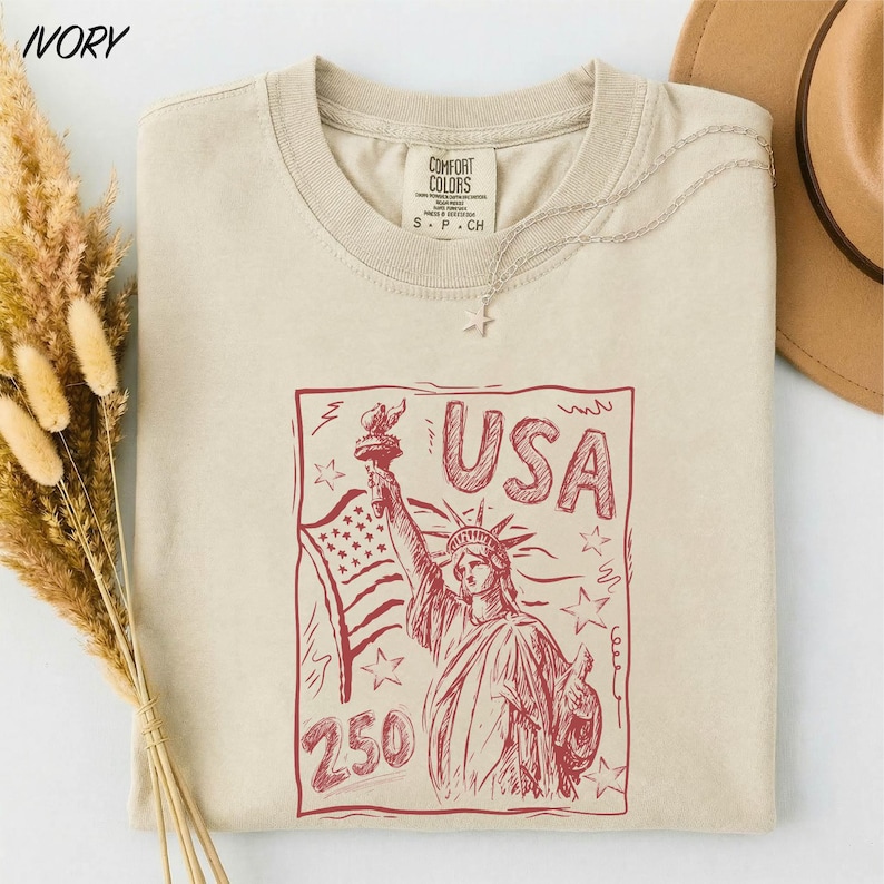 A beige t-shirt with a red graphic design depicting the Statue of Liberty and the text "USA 250" printed on it, along with a necklace and a hat.