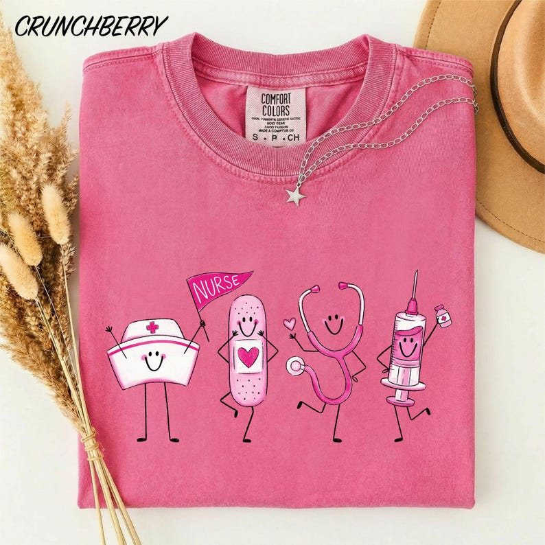 A pink t-shirt with a graphic design featuring cartoon-style illustrations of a nurse, a stethoscope, and a syringe, along with the text "NURSE" and "COMBINED COLORS".