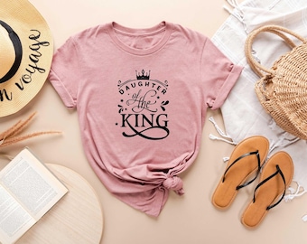 May include: A dusty rose t-shirt with the text "Daughter of the King" in black script and a crown graphic. The shirt is styled with a straw hat, sandals, and a woven bag.