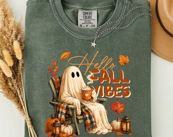 Puede incluir: Una camiseta de cuello redondo color verde salvia con un gráfico de un fantasma sentado en una silla de madera sosteniendo una taza. El texto dice Hello Fall Vibes. El diseño incluye calabazas naranjas, un farol y hojas de otoño. Etiqueta Comfort Colors.