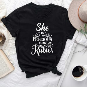 May include: Black t-shirt with the white text "She is far more precious than Rubies." The shirt is styled with a knot at the bottom. A hat, a cup of coffee, and a book are in the background.