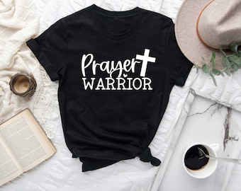 May include: Black t-shirt with the words "Prayer Warrior" and a white cross graphic. The shirt is folded, with a hat, coffee cup, and open book in the background. The shirt is made of a soft material.