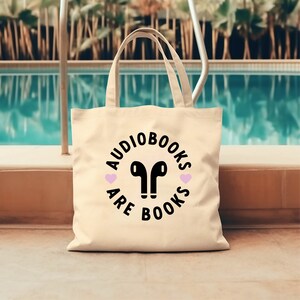 K&ouml;nnte beinhalten: Cremefarbene Tragetasche mit schwarzem Text, der "Audiobooks are Books" in einem kreisf&ouml;rmigen Design zeigt. Das Design umfasst ein Paar schwarze kabellose Ohrh&ouml;rer und zwei rosa Herzen. Die Tasche hat zwei Griffe.