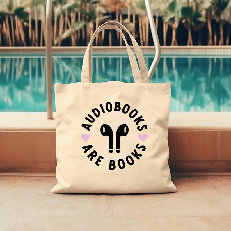 K&ouml;nnte beinhalten: Cremefarbene Tragetasche mit schwarzem Text, der "Audiobooks are Books" in einem kreisf&ouml;rmigen Design zeigt. Das Design umfasst ein Paar schwarze kabellose Ohrh&ouml;rer und zwei rosa Herzen. Die Tasche hat zwei Griffe.