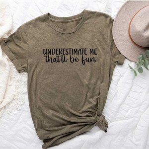 May include: Olive green t-shirt with the text "UNDERESTIMATE ME that'll be fun" in black script. The shirt is knotted at the bottom and is displayed on a white surface. A beige hat with a brown band is visible.