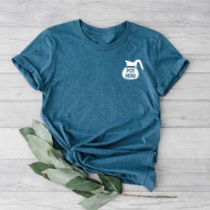 May include: A blue t-shirt with a white graphic of a coffee pot and the words "POT HEAD" printed on the front. The shirt is made of a soft material and has rolled-up sleeves. Green leaves are placed at the bottom of the shirt.