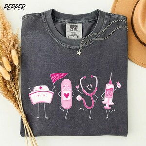 A gray t-shirt with a graphic design featuring cartoon-style illustrations of a nurse, a stethoscope, a syringe, and a heart, along with the text "NURSE" and "COMFORT COLORS".