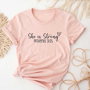 May include: A light pink t-shirt with the text "She is Strong" and "Proverbs 31:25" in black script. A small heart is next to the word "Strong". The shirt has rolled-up sleeves and is styled on a neutral background.