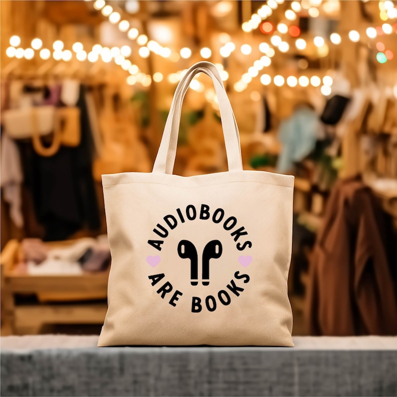 K&ouml;nnte beinhalten: Beige Tragetasche mit dem Text "AUDIOBOOKS ARE BOOKS" in einem schwarzen Kreisdesign, mit schwarzen Kopfh&ouml;rer-Symbolen und rosa Herz-Akzenten. Die Tasche hat zwei Tragegriffe.