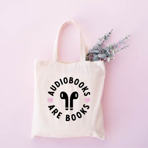 K&ouml;nnte beinhalten: Eine cremefarbene Tragetasche mit dem Schriftzug "Audiobooks are Books" in einem Kreis, mit schwarzen Kopfh&ouml;rern und rosa Herzdetails. Die Tasche ist aus hellem Stoff gefertigt und hat Henkel. Ein Zweig mit Gr&uuml;n ist sichtbar.