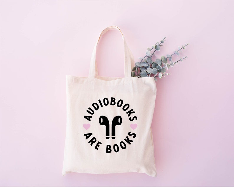 K&ouml;nnte beinhalten: Eine cremefarbene Tragetasche mit dem Schriftzug "Audiobooks are Books" in einem Kreis, mit schwarzen Kopfh&ouml;rern und rosa Herzdetails. Die Tasche ist aus hellem Stoff gefertigt und hat Henkel. Ein Zweig mit Gr&uuml;n ist sichtbar.