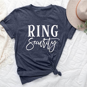 May include: A dark gray t-shirt with the words "RING Security" printed in white. The shirt is knotted at the bottom and is laying on a white surface. A beige hat with a brown band is in the background.