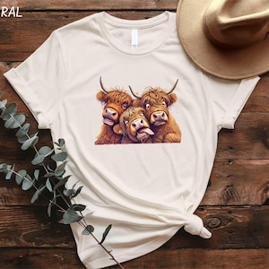A white t-shirt with a graphic of three yaks sticking their tongues out, along with a beige hat and some greenery, all placed on a wooden surface.