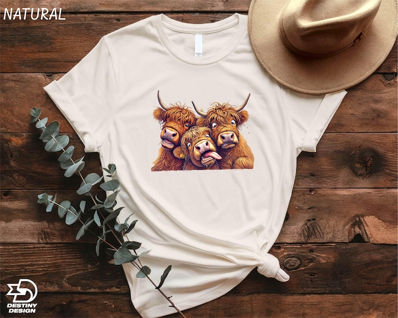 A white t-shirt with a graphic of three yaks sticking their tongues out, along with a beige hat and some greenery, all placed on a wooden surface.