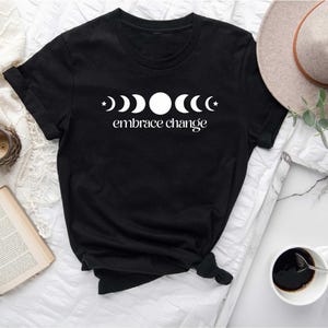 May include: Black t-shirt featuring a white graphic of moon phases and the words "embrace change". The shirt is short-sleeved and has a crew neck. A neutral-colored hat and a cup of coffee are in the background.