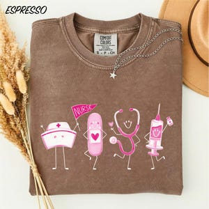 a brown t-shirt with a design of four cartoon-style medical professionals, including a nurse, a stethoscope, a syringe, and a pill bottle, all depicted in a playful and colorful manner.
