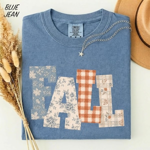 May include: A blue short-sleeve t-shirt with the word FALL printed in large block letters. Each letter features a different pattern: blue floral, cream floral, orange and white gingham, and a cream plaid with small orange pumpkin motifs.