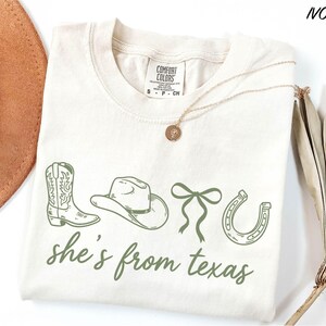 a white t-shirt with a graphic design of a cowboy hat, a horseshoe, and a ribbon, along with the text "she's from texas" printed on it. The t-shirt is laid out on a white surface, with a brown hat and a dried plant visible in the background.