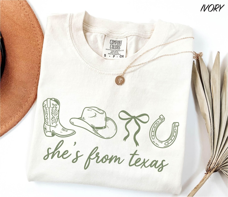 a white t-shirt with a graphic design of a cowboy hat, a horseshoe, and a ribbon, along with the text "she's from texas" printed on it. The t-shirt is laid out on a white surface, with a brown hat and a dried plant visible in the background.