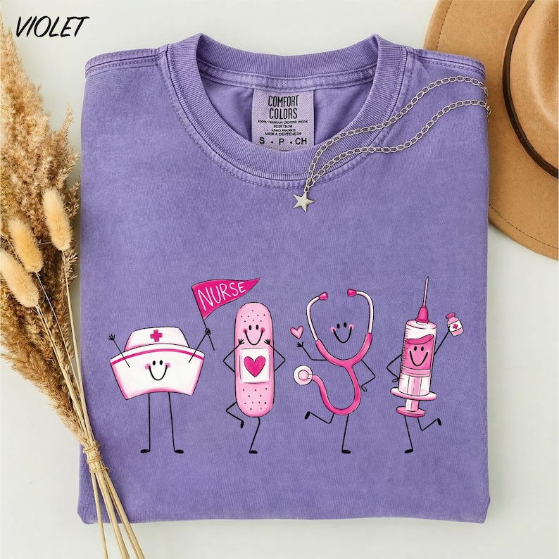 a purple t-shirt with a design of four cartoon nurse characters, including a nurse's hat, bandages, a stethoscope, and a syringe, all smiling and dancing.