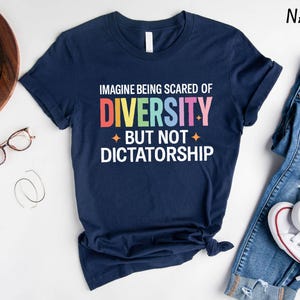 May include: Navy blue t-shirt with the text "IMAGINE BEING SCARED OF DIVERSITY BUT NOT DICTATORSHIP" in a rainbow color scheme. The shirt is styled with a pair of blue jeans and white sneakers.