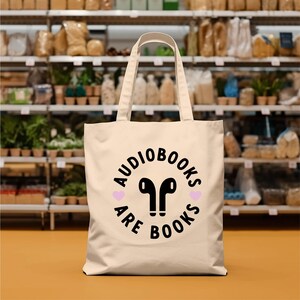 K&ouml;nnte beinhalten: Cremefarbene Tragetasche mit dem Text "AUDIOBOOKS ARE BOOKS" in einem schwarzen Kreisdesign, mit schwarzen Ohrh&ouml;rern und zwei rosa Herzen. Die Tasche hat eine rechteckige Form mit zwei Griffen.
