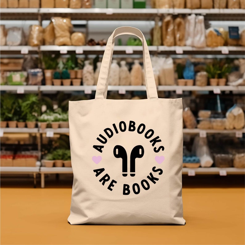 K&ouml;nnte beinhalten: Cremefarbene Tragetasche mit dem Text "AUDIOBOOKS ARE BOOKS" in einem schwarzen Kreisdesign, mit schwarzen Ohrh&ouml;rern und zwei rosa Herzen. Die Tasche hat eine rechteckige Form mit zwei Griffen.