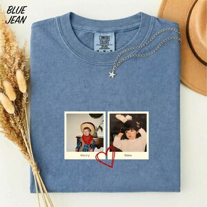 A blue t-shirt with a photo of two children, Henry and Emma, printed on it, along with a heart symbol. The t-shirt is placed on a white surface, and there is a brown hat and some dried flowers nearby.