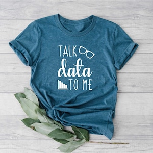 May include: A teal blue t-shirt with the phrase "TALK data TO ME" in white script. The design includes glasses and a bar graph icon. The shirt is folded and has a casual, relaxed fit.