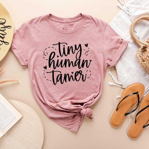 May include: A dusty rose t-shirt with the text "tiny human tamer" in black script. The shirt is knotted at the bottom and is next to a straw hat, a woven bag, a pair of brown sandals, and an open book.