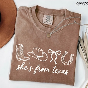 a brown t-shirt with the text "she's from texas" printed on it, along with various western-themed icons such as a cowboy hat, a boot, and a horseshoe. The t-shirt is laid out on a white surface, and there is a brown hat and some dried leaves nearby.